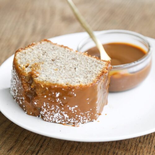 Slice of banana loaf cake with a dip of dulce de leche on a white plate on a wood surface.