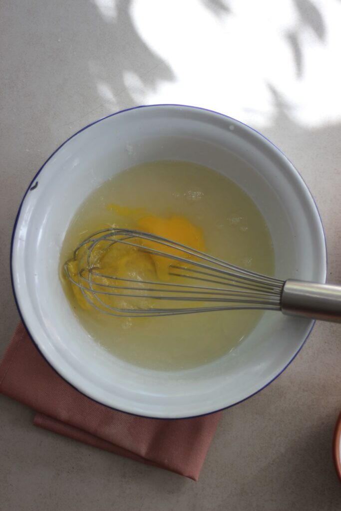 A bowl with egg yolk and a liquid, and a hand whisk.