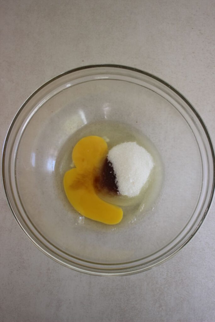 A glass bowl with sugar, whites, and egg yolk on a white surface.