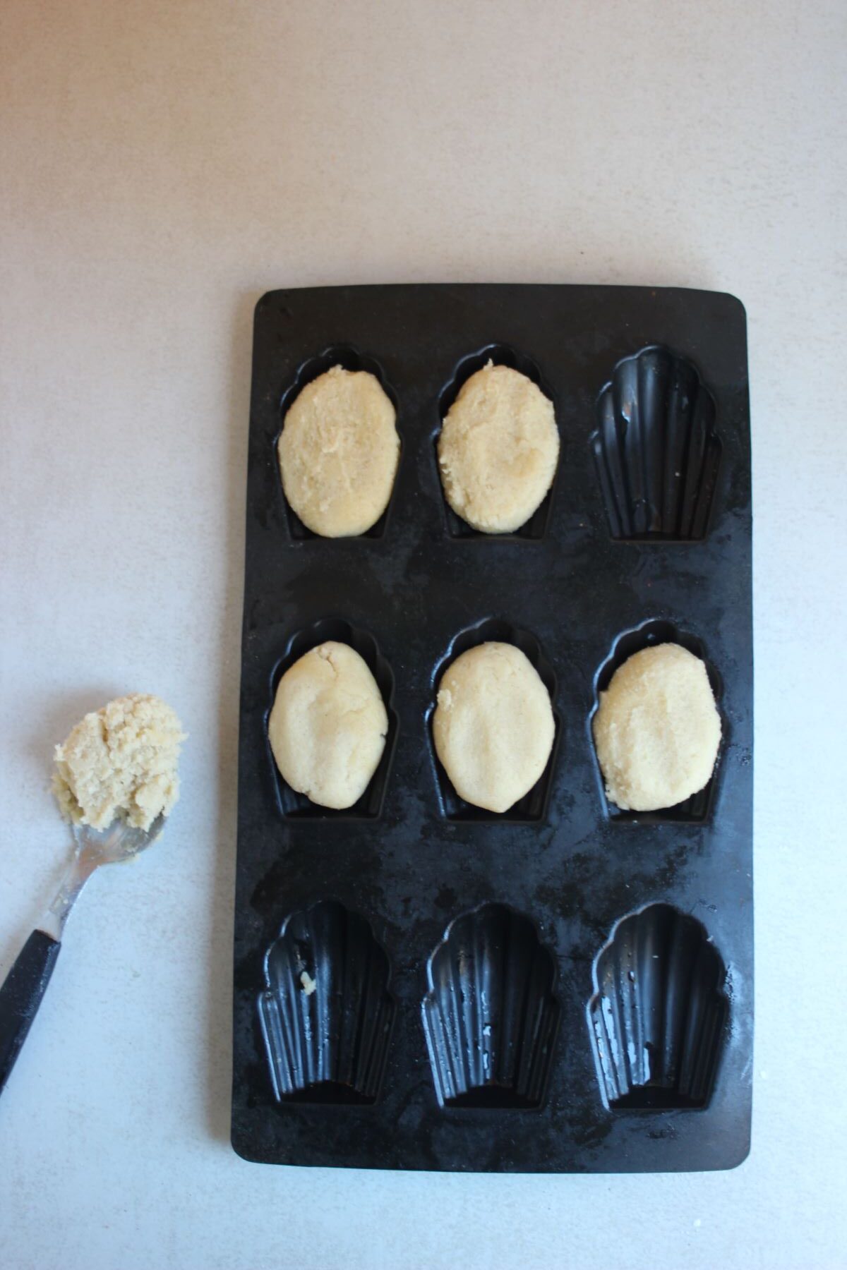 Silicone madeleine pan, some holes with dough, and a spoon with dough on the side.