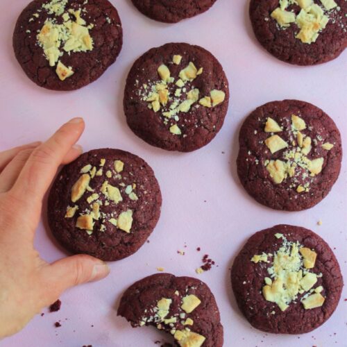 Red velvet cookies on a pink surface and a hand about to grab one.