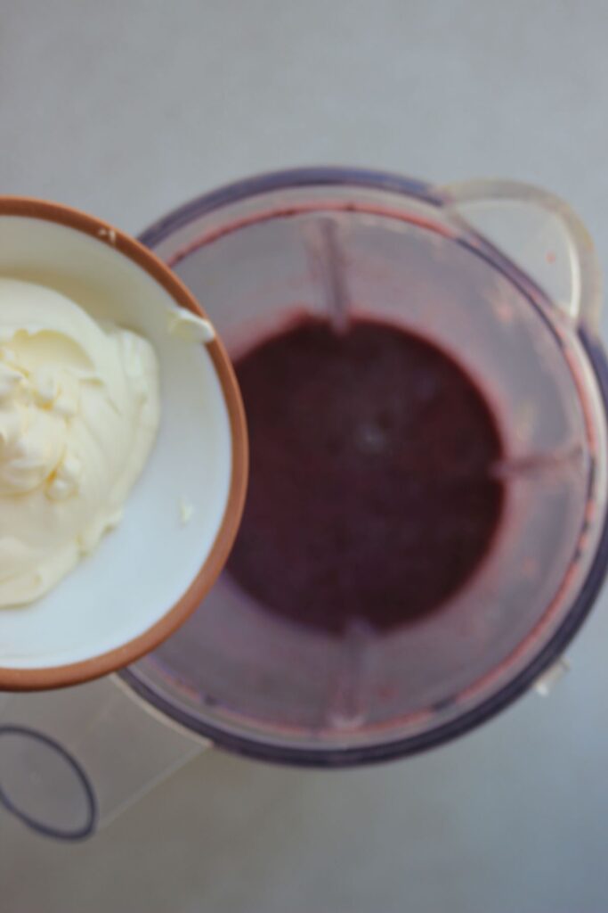 Bowl with cream is about to be poured into the blender with a violet liquid.