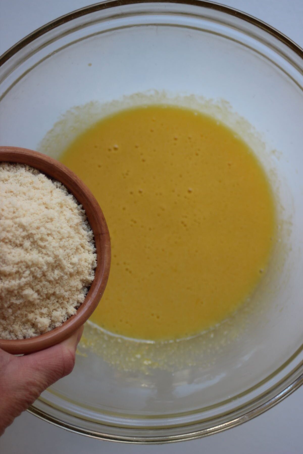Bowl with almond flour is about to be poured into glass bowl with yellow mixture.