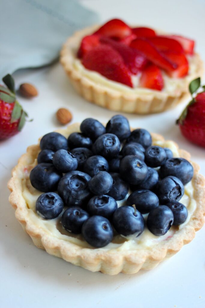 Vanilla pastry cream and fresh blueberry tartlet on a white surface. Strawberry tartlet behind, and fresh strawberries on the sides.
