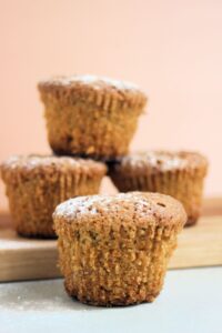 A carrot cake muffin on a white surface, more muffins behind on a wooden board.