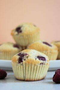 Raspberry muffin and raspberries on the sides. More muffins behind on a white plate.
