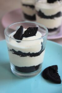 Glass with lemon mousse and Oreos on a light blue plate. Broken Oreos on the side.