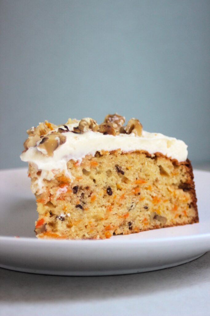 Portion of carrot cake with cream cheese frosting and chopped walnuts on an white plate.