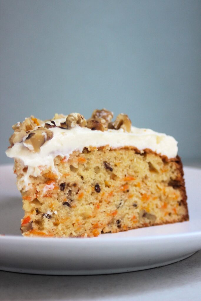 Portion of carrot cake with cream cheese frosting and chopped walnuts on an white plate.