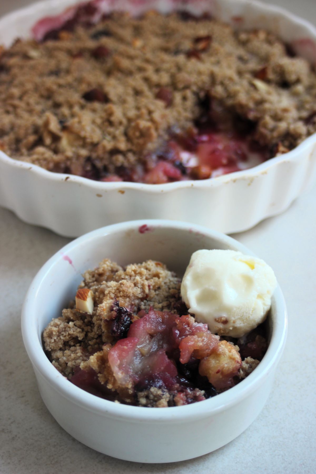 Small bowl with apple crumble and a scoop of ice cream. A baking dish with apple crumble behind.