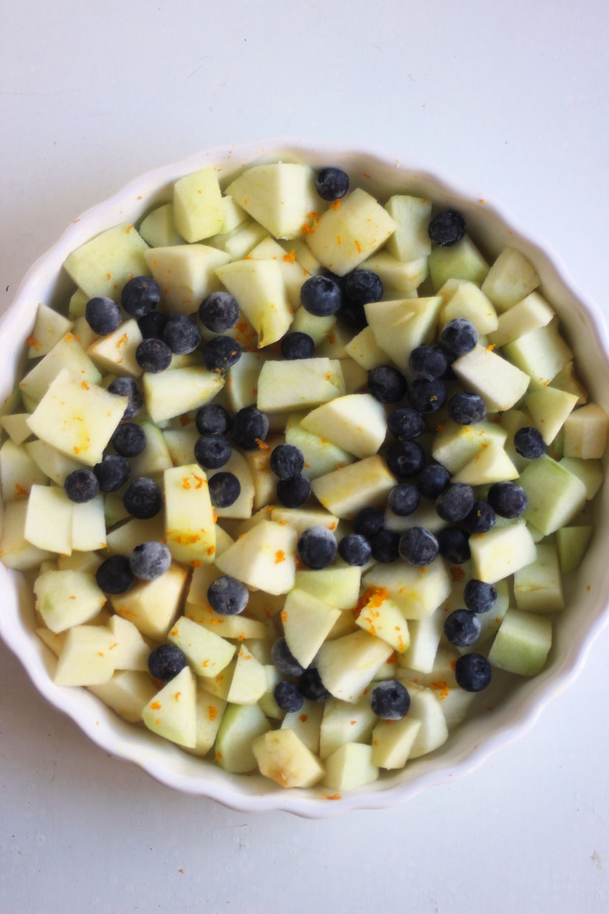 Round white baking dish with peeled and chopped apples, and blueberries seen from above.