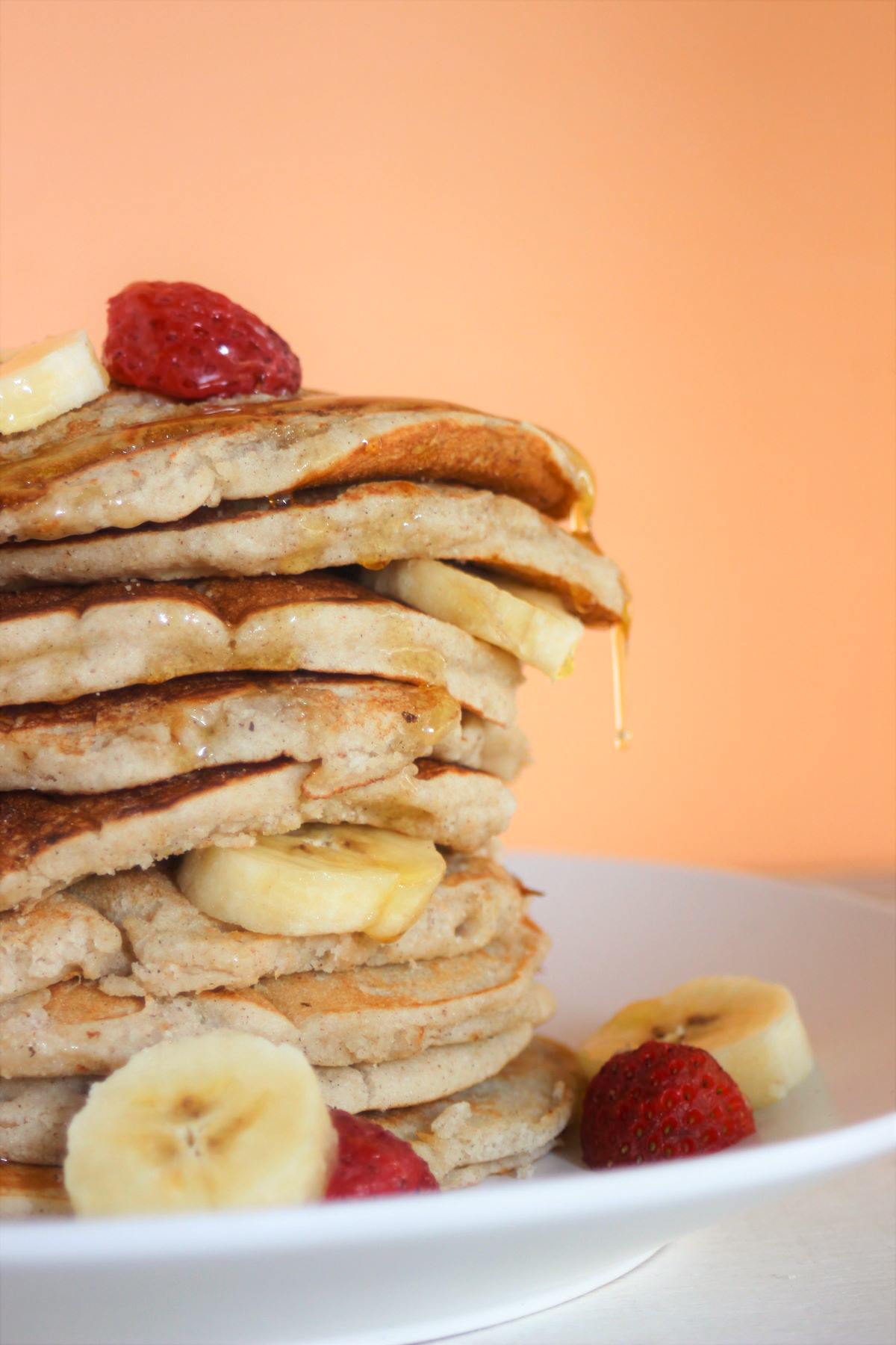 Tower of banana pancakes with banana slices and strawberries on a white plate. Light orange background.