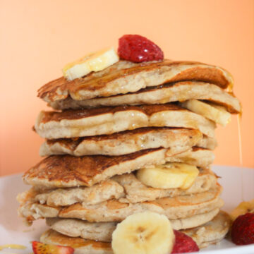 Tower of banana pancakes with banana slices and strawberries on a white plate. Orange background.