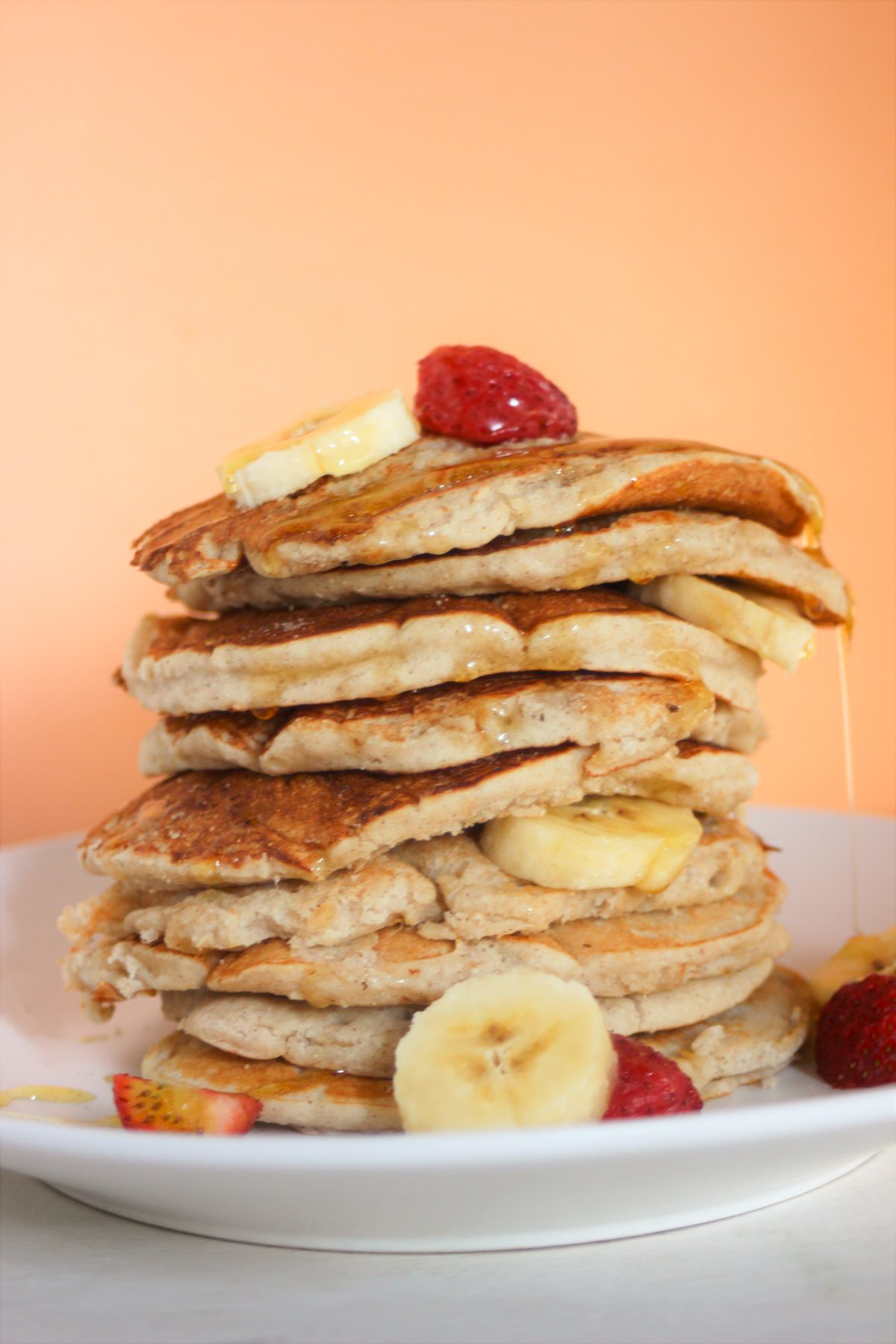 Tower of banana pancakes with banana slices and strawberries on a white plate. Orange background.