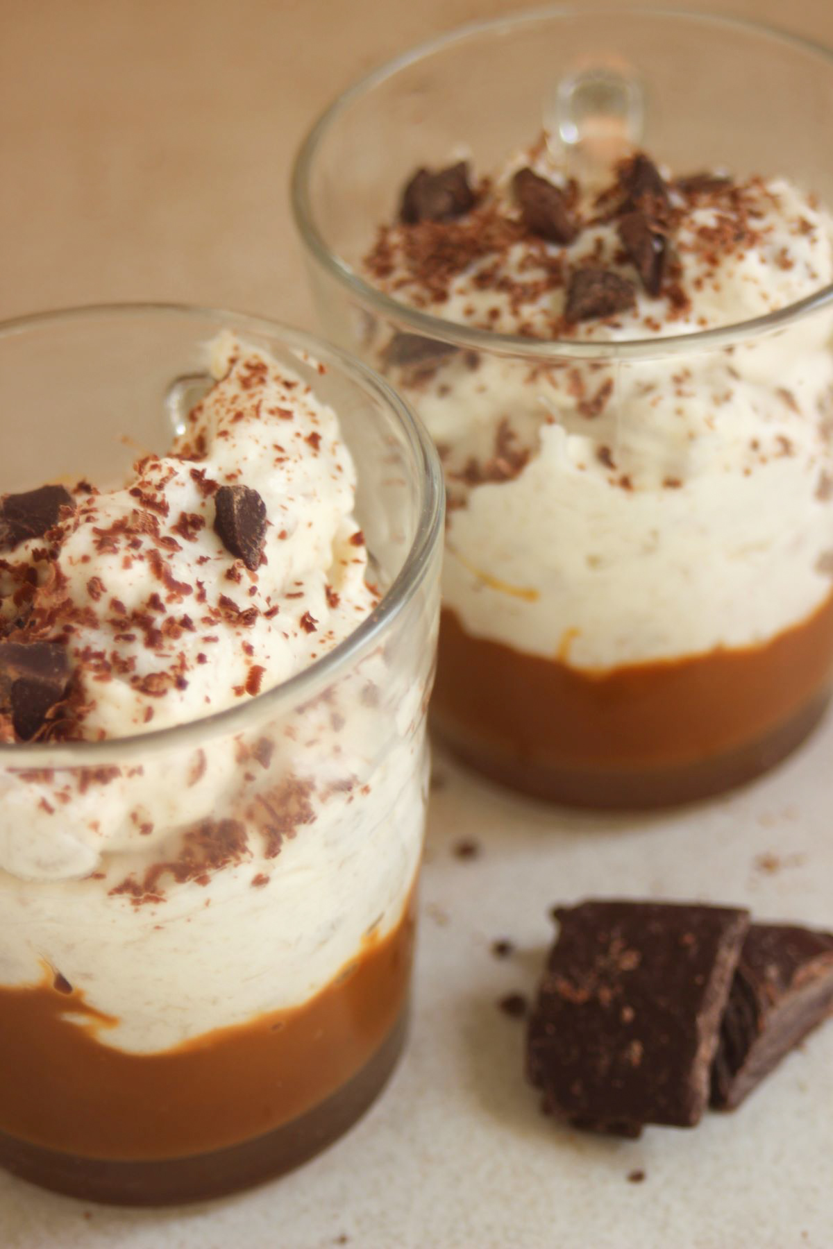 Two glasses with dulce de leche, banana cream, and shredded chocolate. Chocolate chunks on the surface.