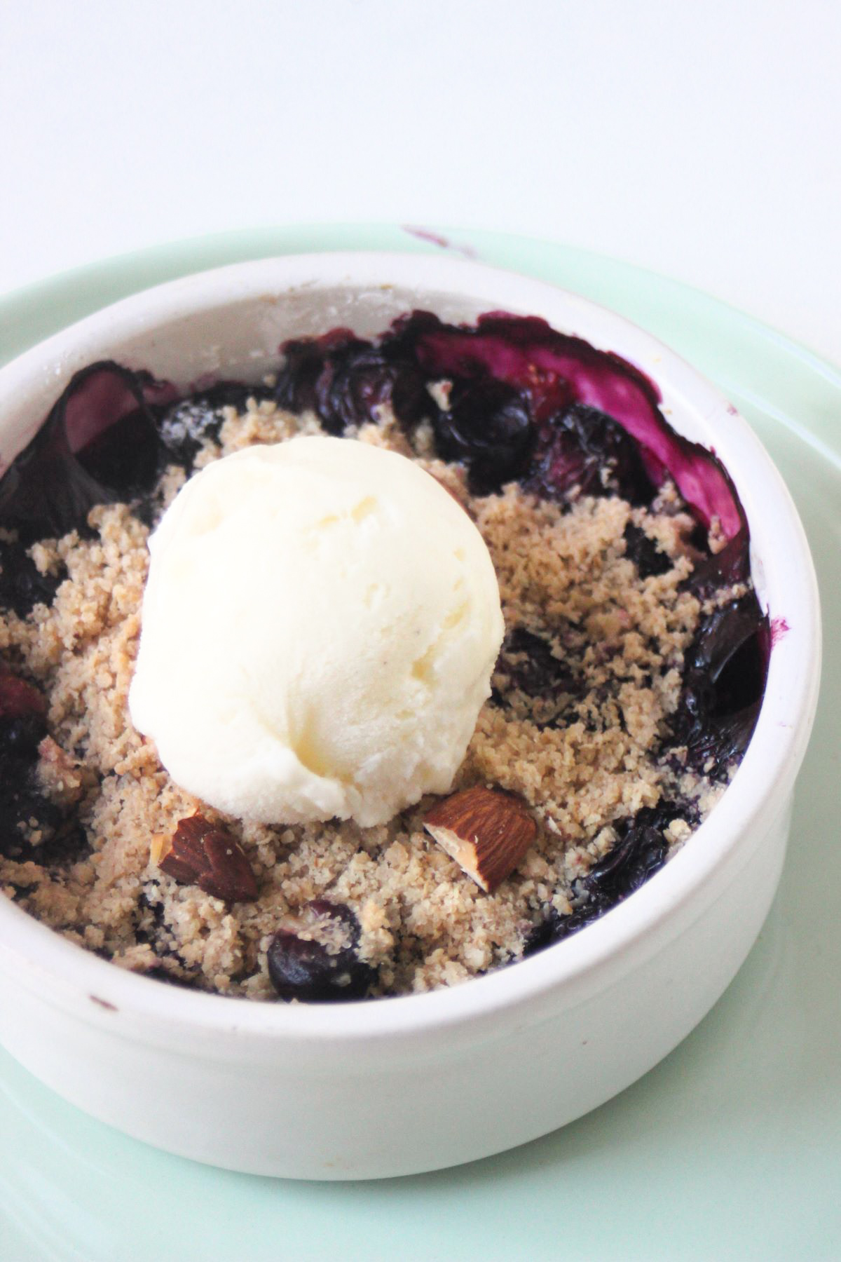 Blueberry crisp and a scoop od ice cream in a small round white bowl on a aqua green plate.