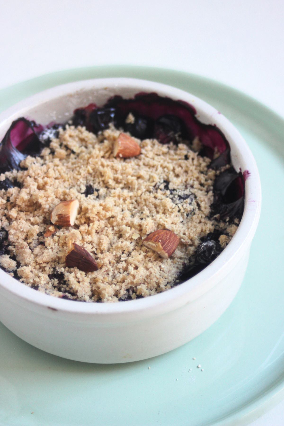 Blueberry crisp in a small round white bowl on a aqua green plate.