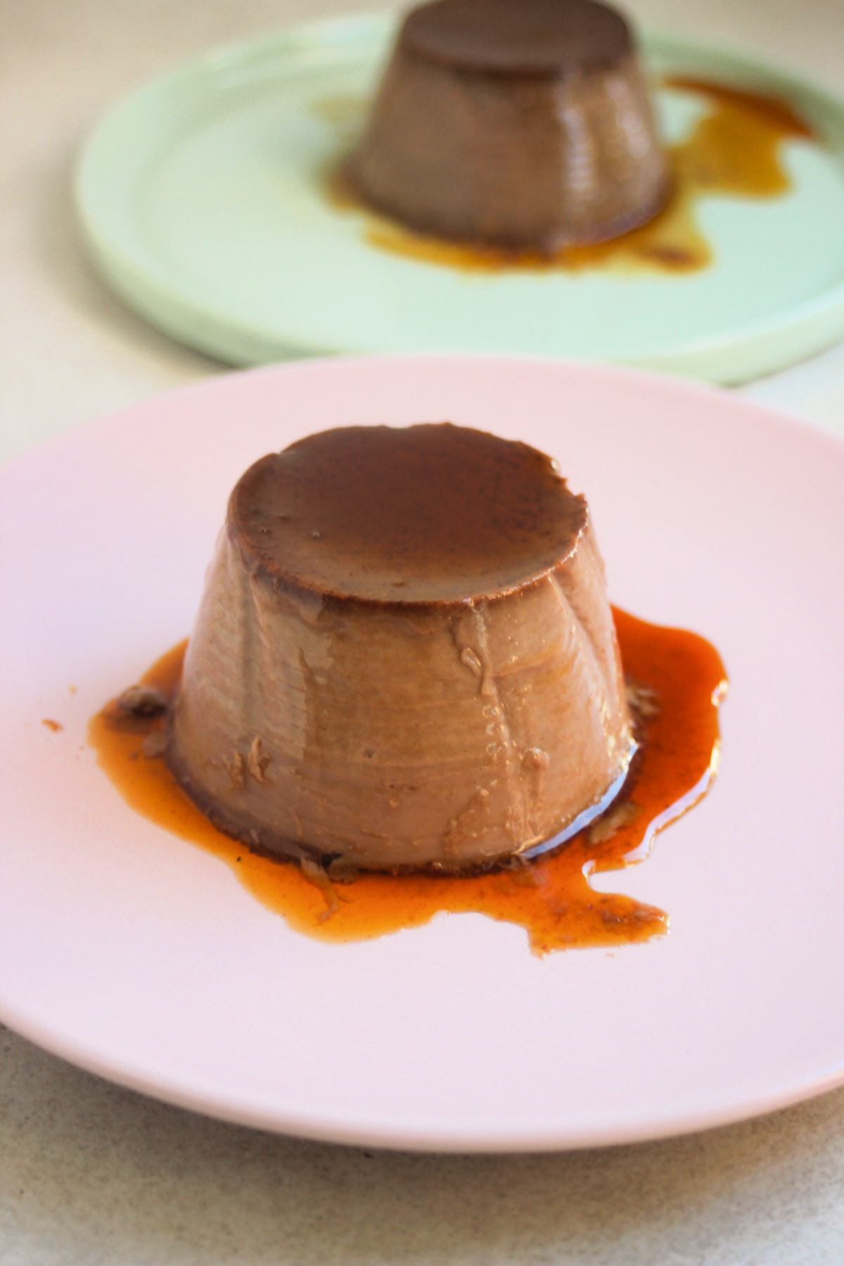 Chocolate flan with caramel on a pink plate. Behind another chocolate flan on an aqua-green