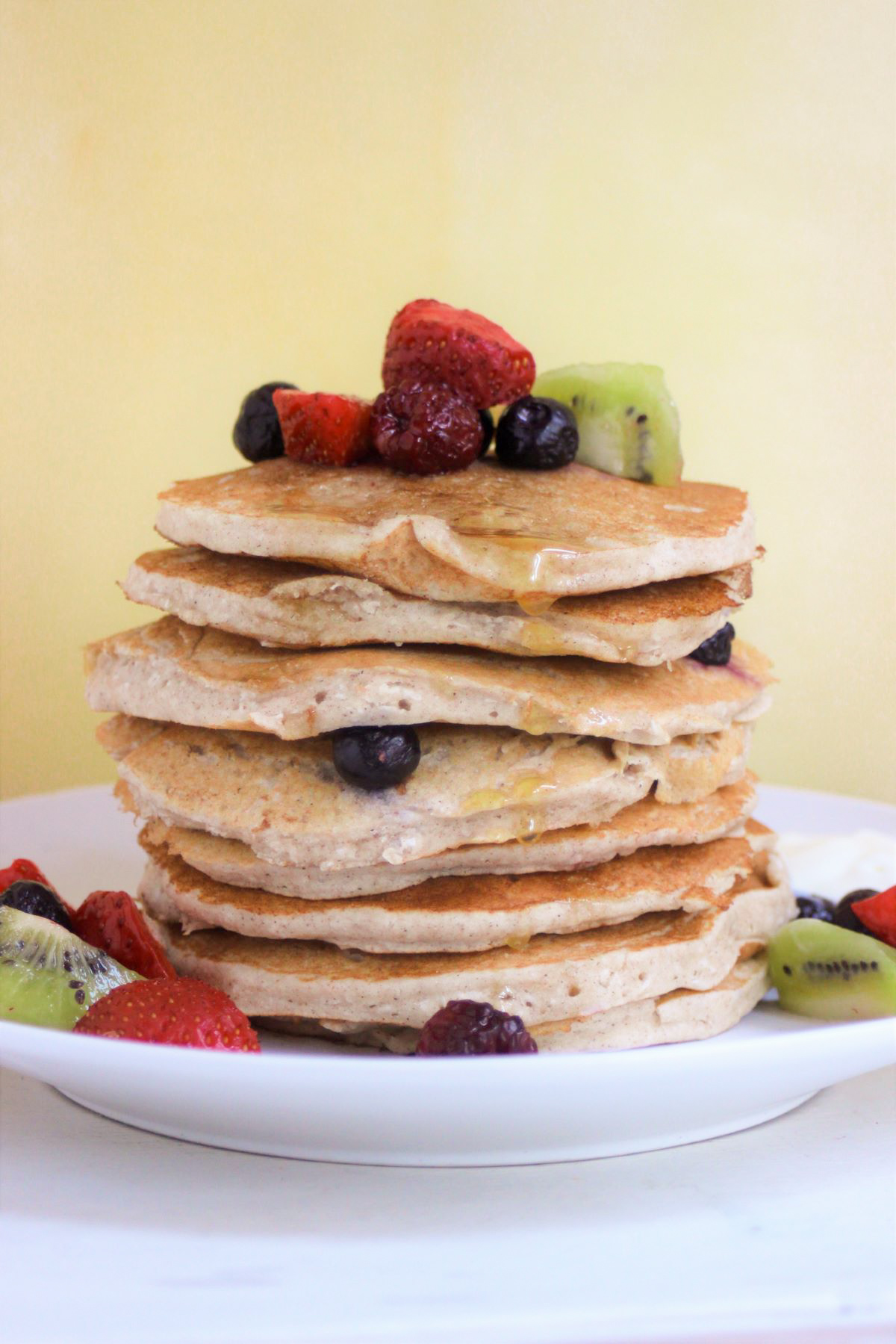 A tower of pancakes with strawberries, blueberries, and kiwis on a white plate. Yellow background.