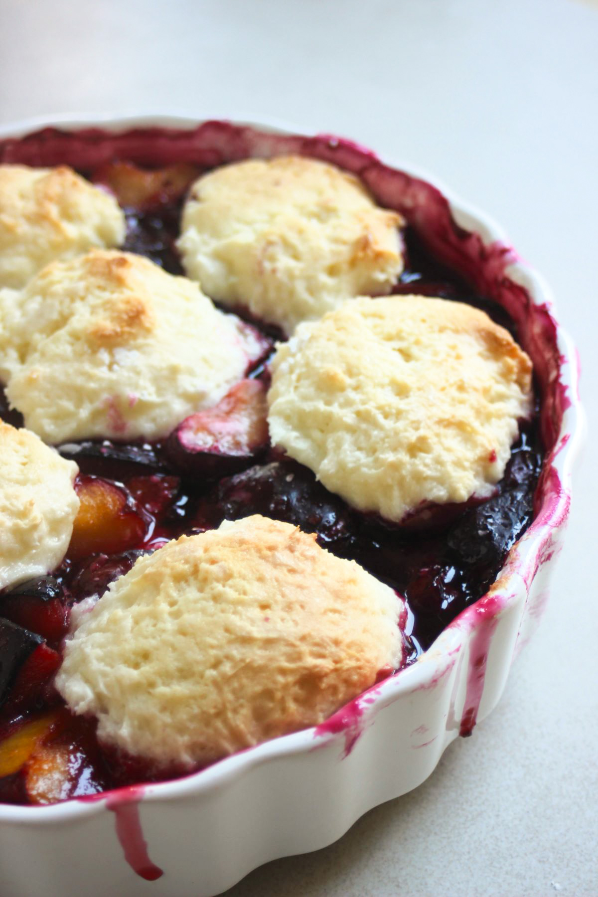 Round white baking dish with plum cobbler on a white surface.