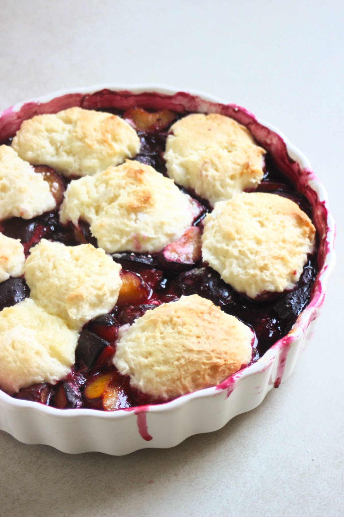 Round white baking dish with plum cobbler on a white surface seen from above.