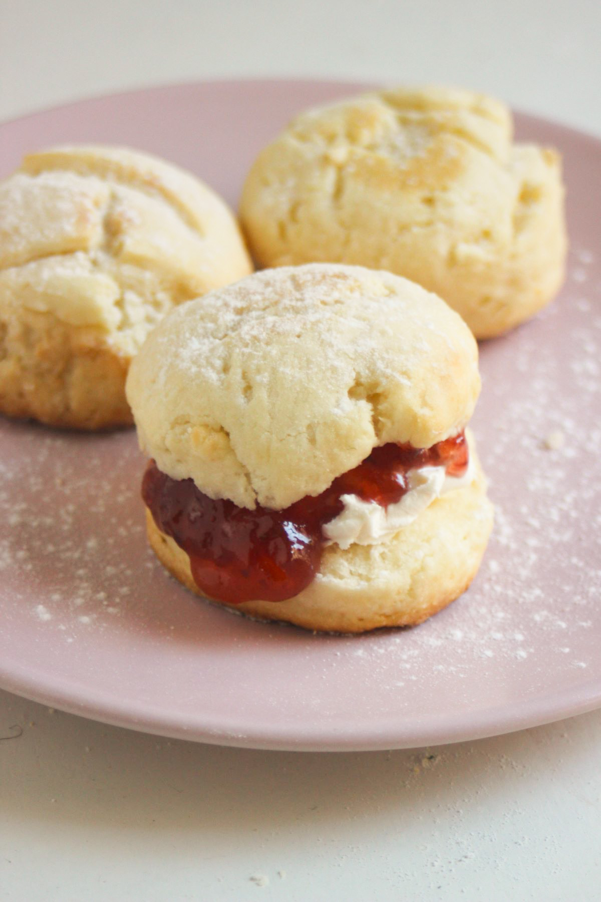 Scon filled with cream cheese and jam on a pink plate. More scones behind.