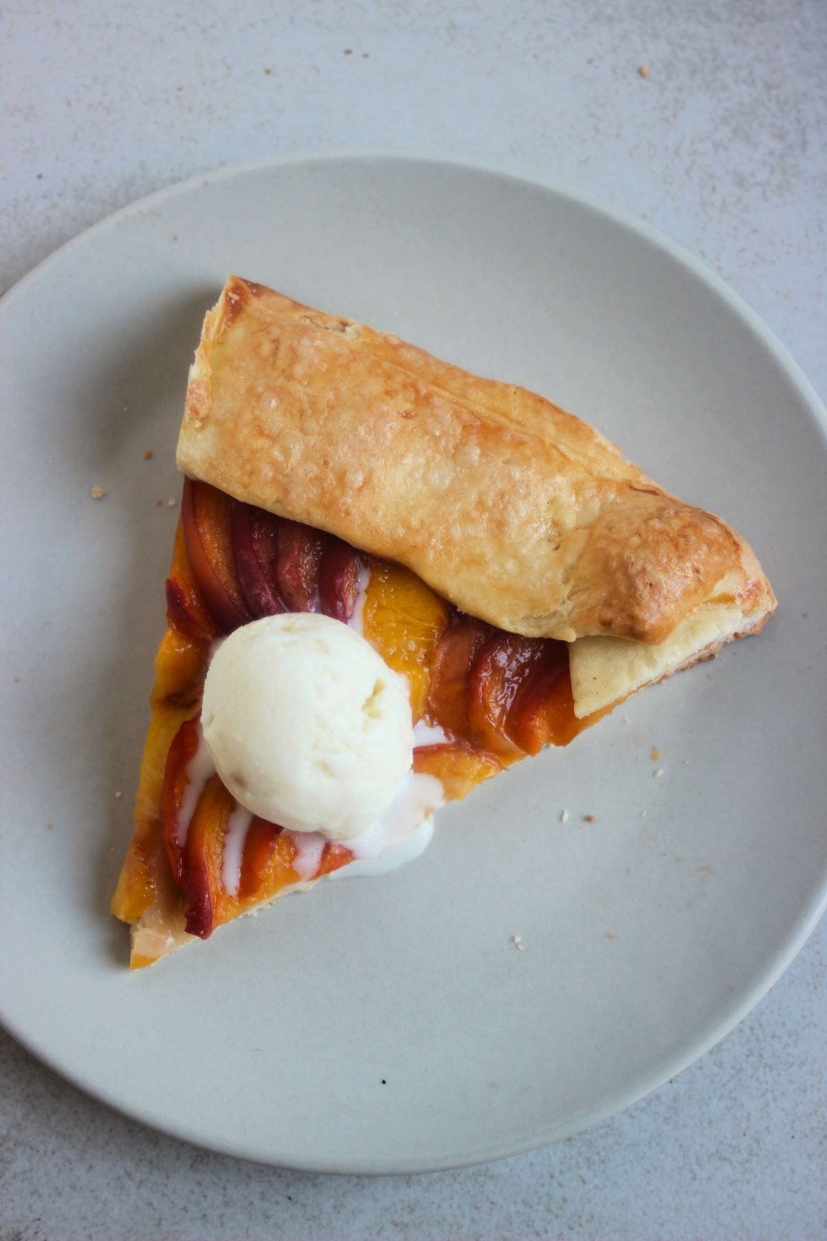 Portion of a peach galette with a scoop of ice cream on a light grey plate seen from above.