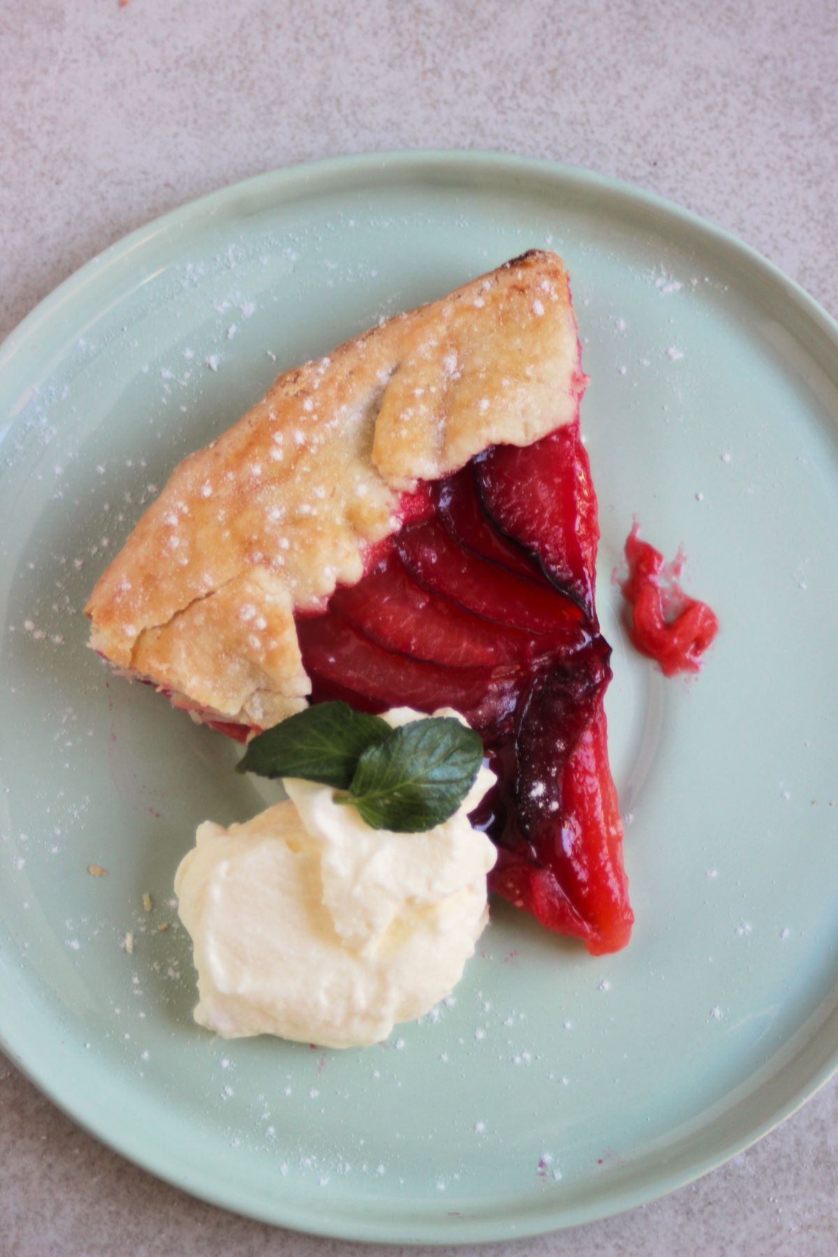 Portion of plum tart with whipped cream and two mint leaves on an aqua-green plate seen from above.