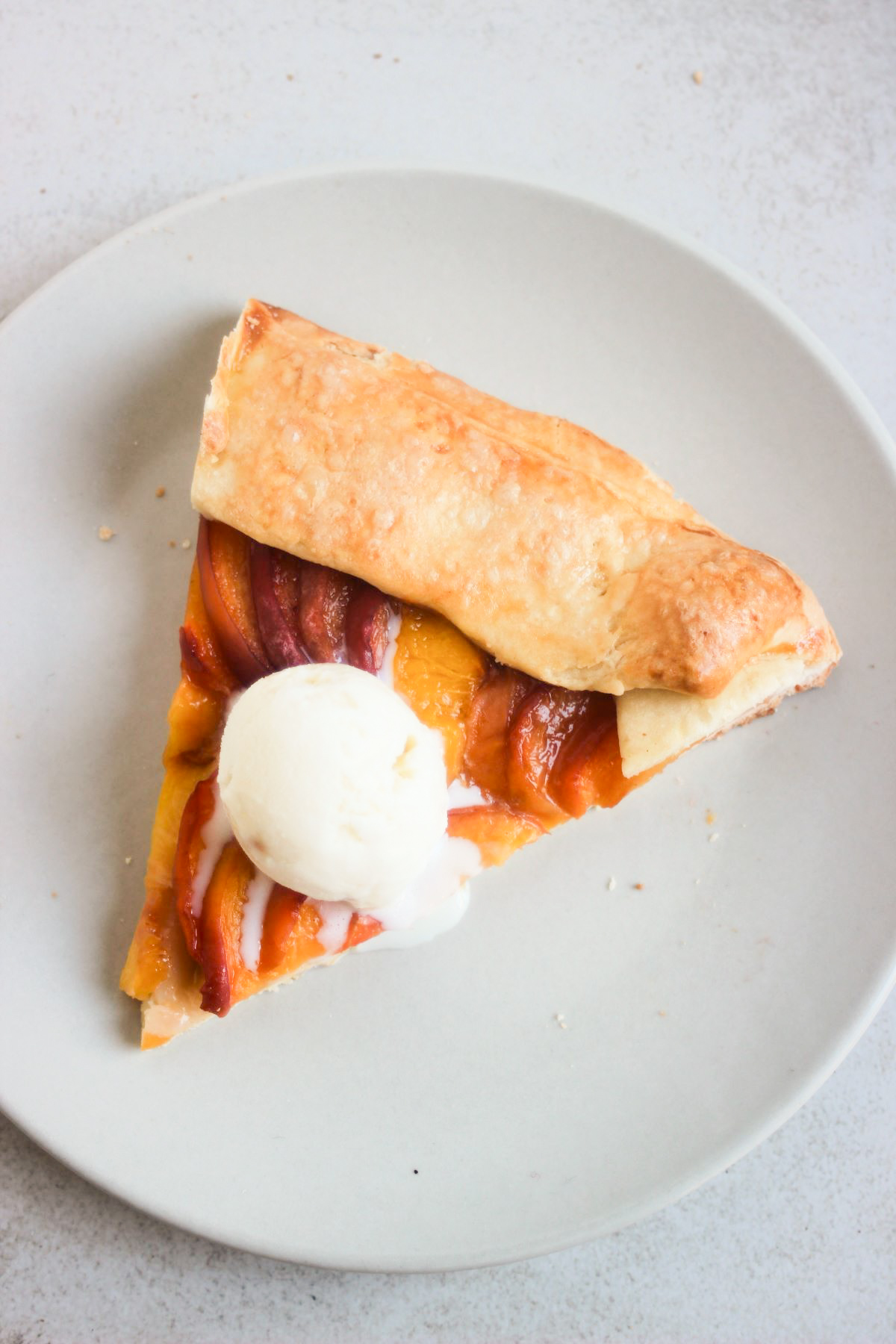 Portion of a peach galette with a scoop of ice cream on a light grey plate seen from above.