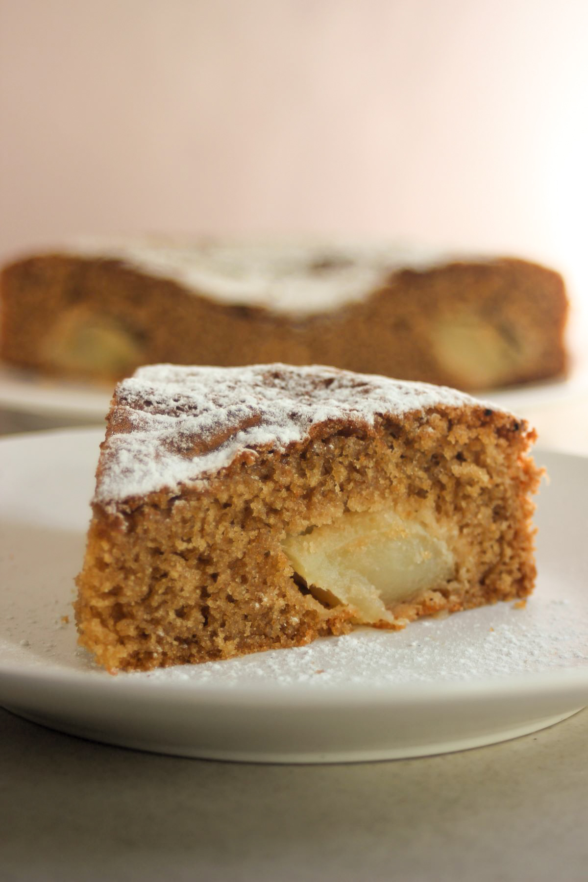 A portion of apple cake on a white plate. The cake behind.