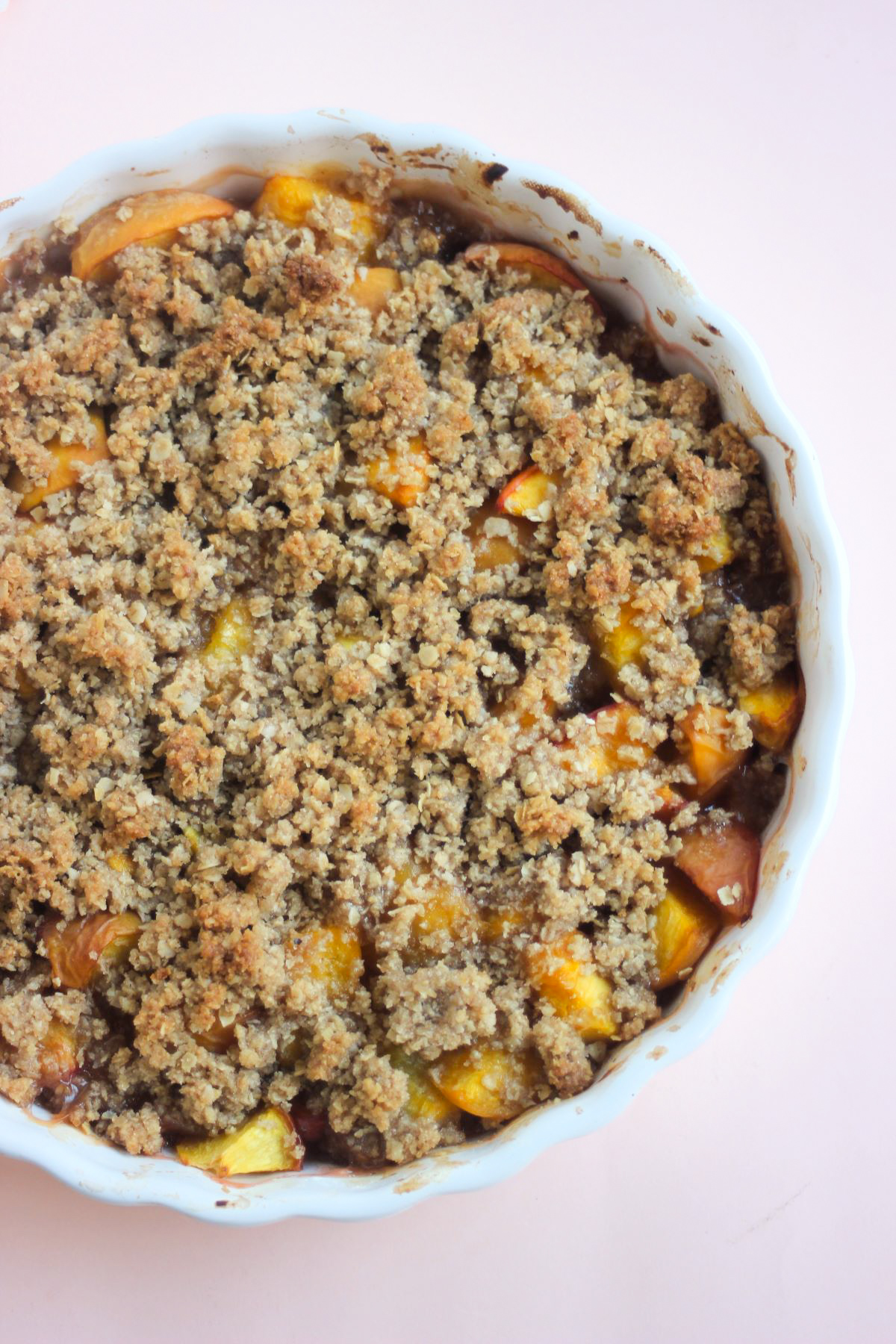 White round baking dish with peach crisp on a pink surface seen from above.