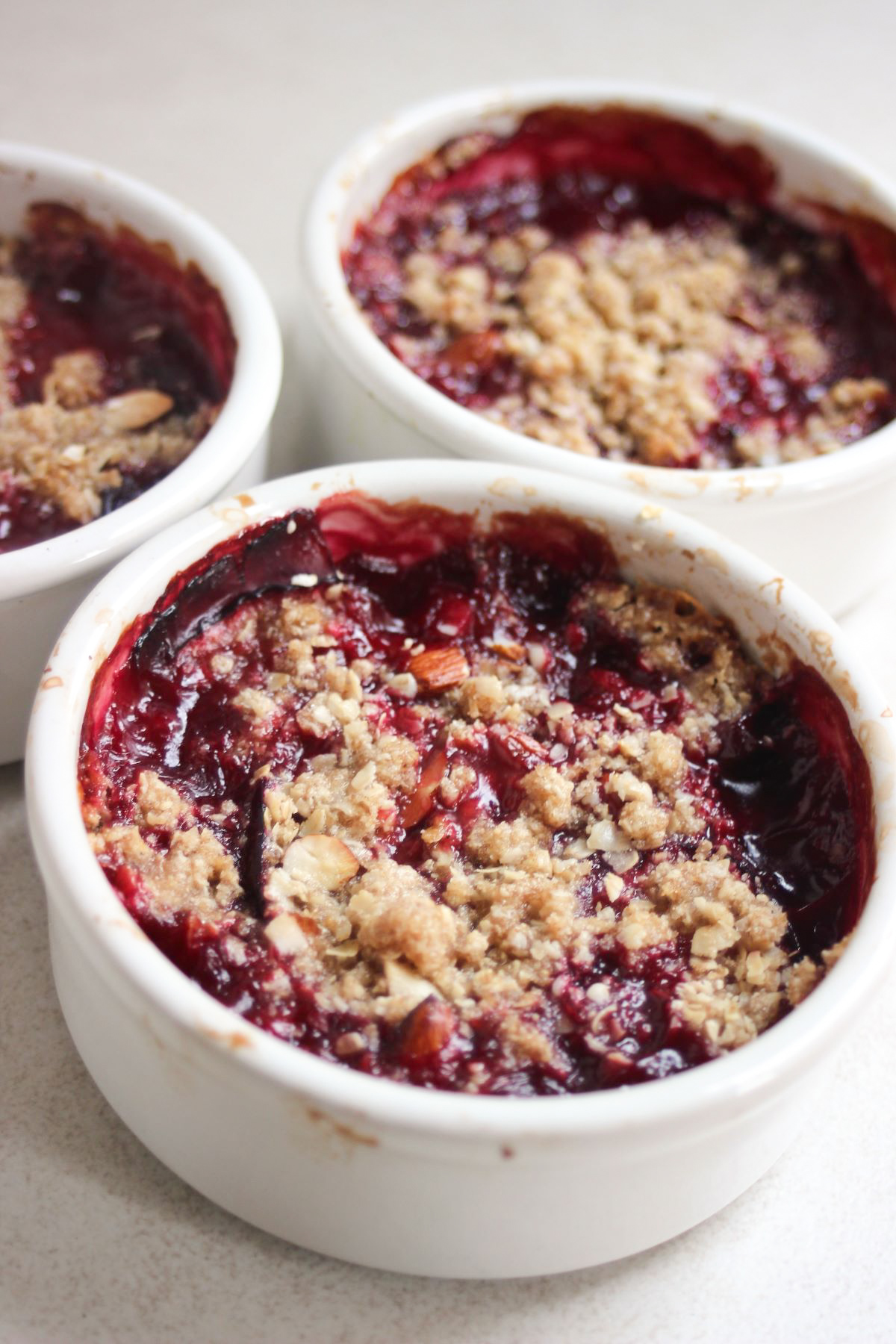 Three ramekins with plum crisp on a white surface.