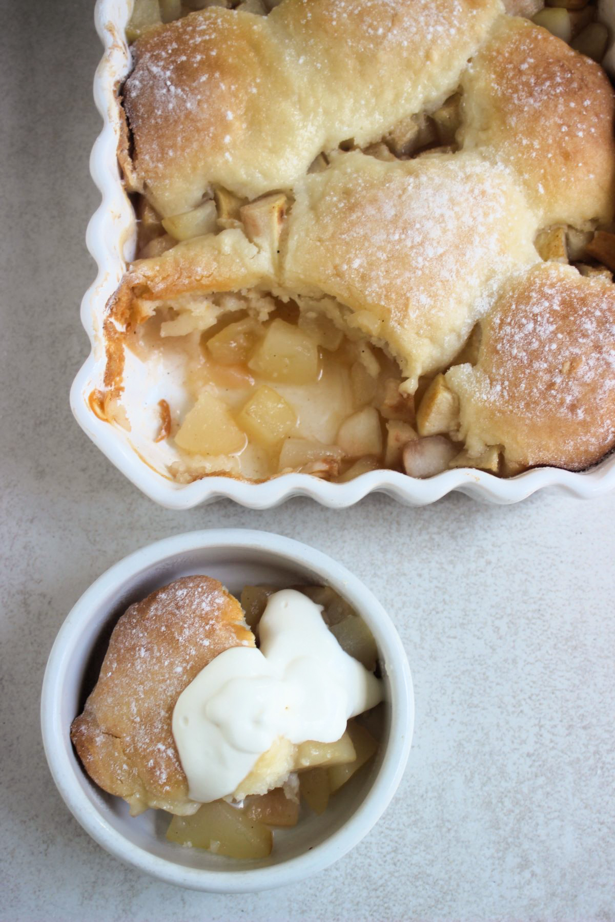 Rectangular white baking dish with pear cobbler without a portion and small bowl with pear cobbler with whipped cream seen from above.