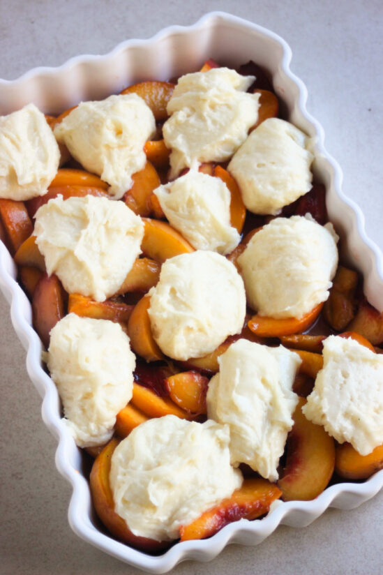 Rectangular baking dish with sliced peaches and scoops of batter.