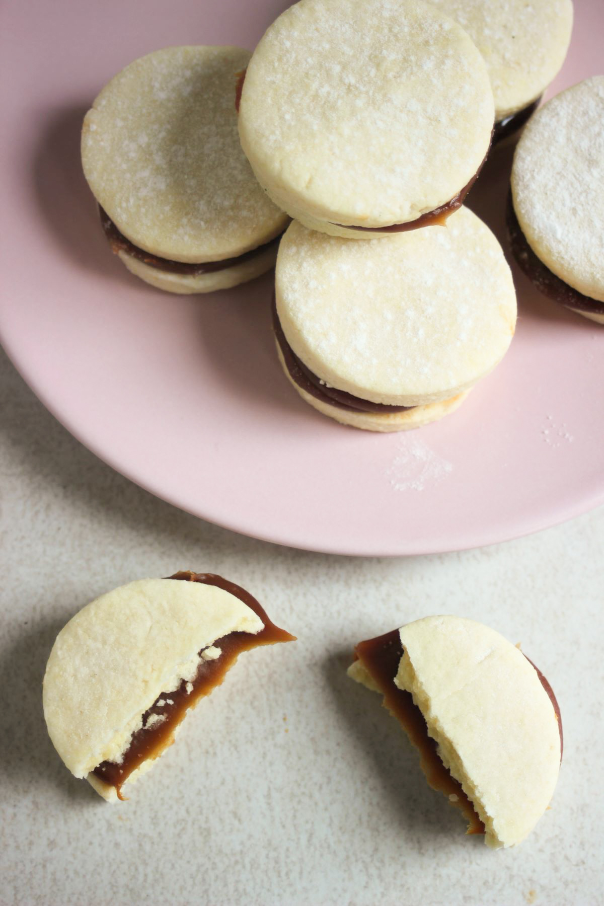 Alfajores on a pink plate and one alfajor cut in half ahead of the plate. Everithing seen from above.