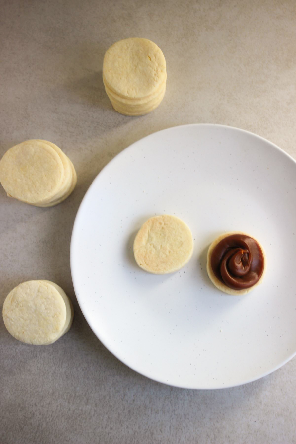 Two biscuits of alfajor dough, one with dulce de leche, on a white plate. Many biscuits of alfajor dough on the side.