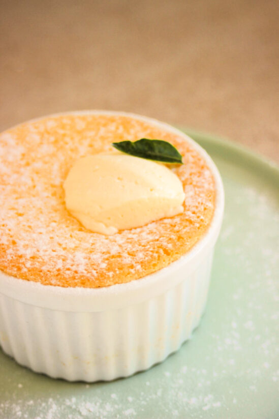 White ramekin with lemon pudding cake and whipped cream on an aqua green plate.