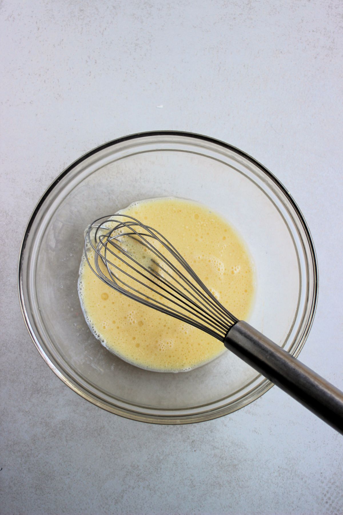 Glass bowl with a light yellow liquid and a hand whisk.