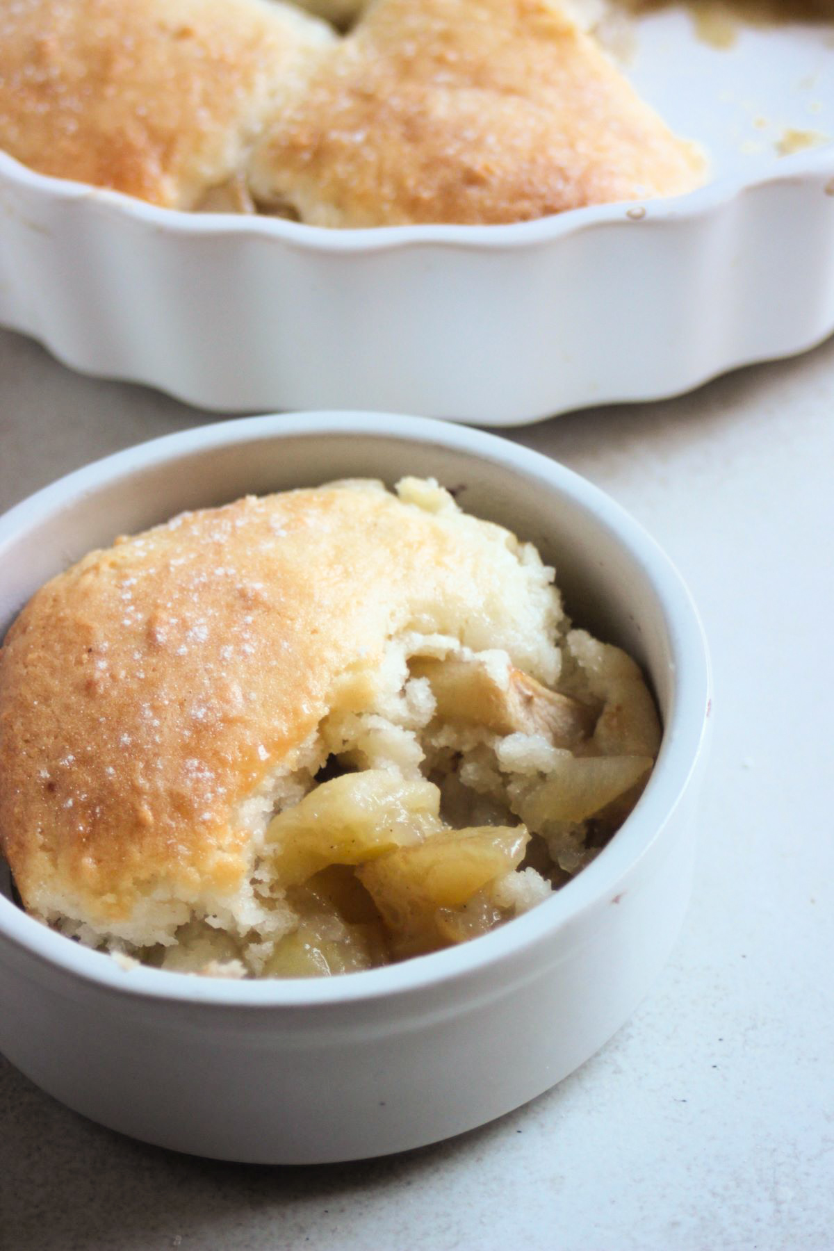 Small bowl with a portion of apple cobbler. Apple cobbler behind.