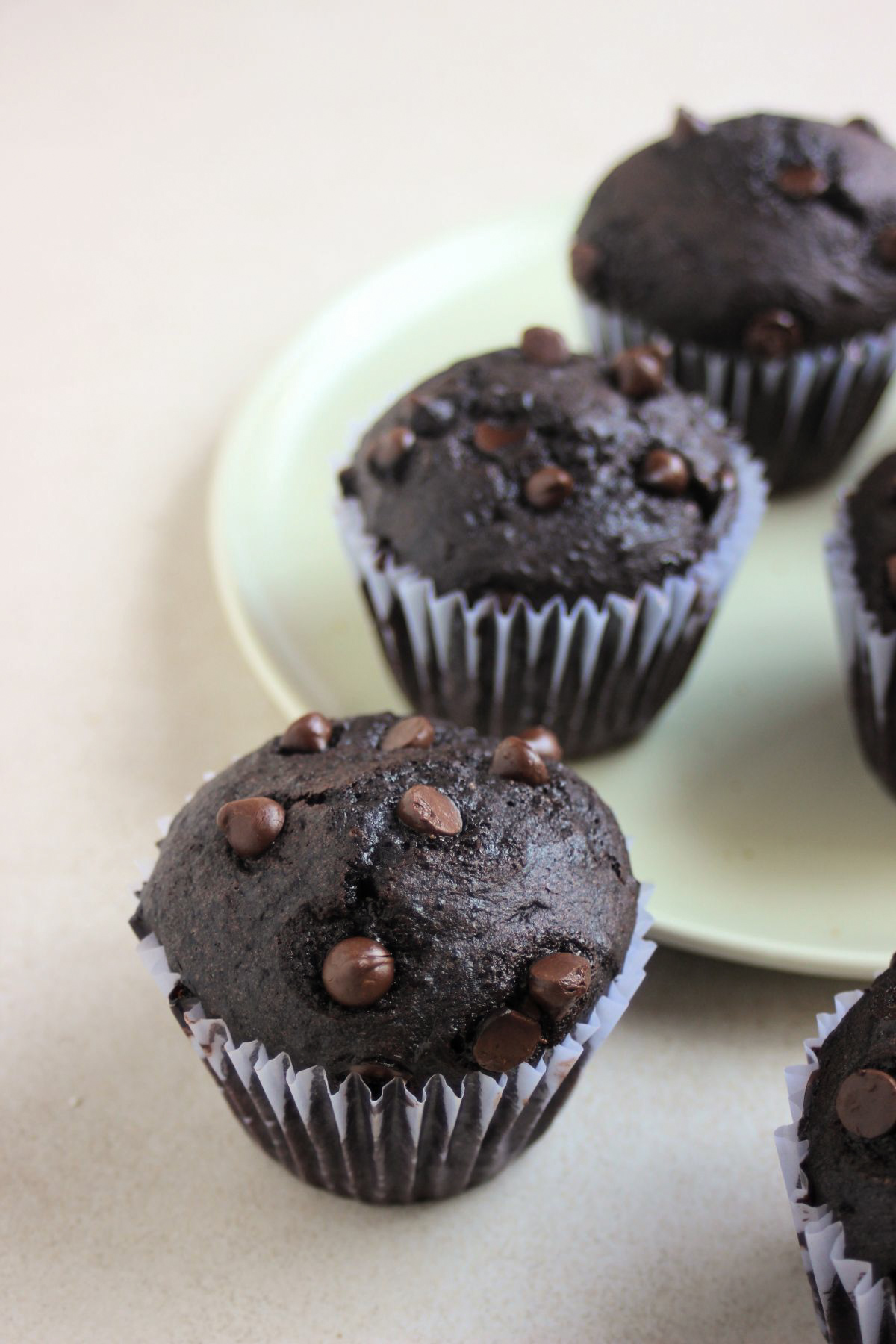 Double chocolate chip muffin with a liner on a white surface. More muffins behind on an aqua green plate.