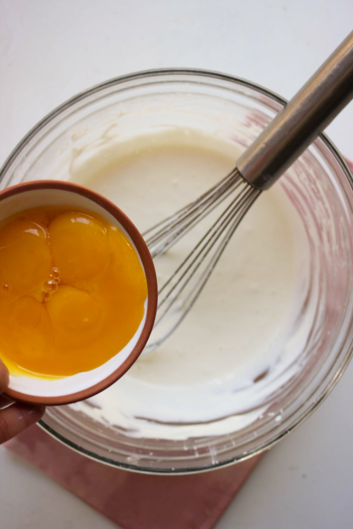 Small bowl with egg yolks is about to be poured into a glass bowl with a white mixture and a hand whisk.