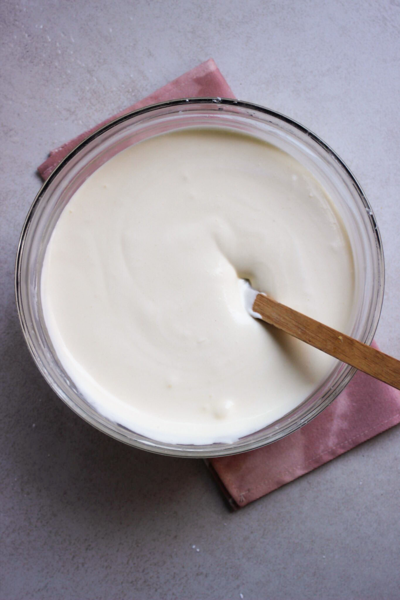 Glass bowl with a white and smooth mixture and a spatula. Pink napkin under the bowl.