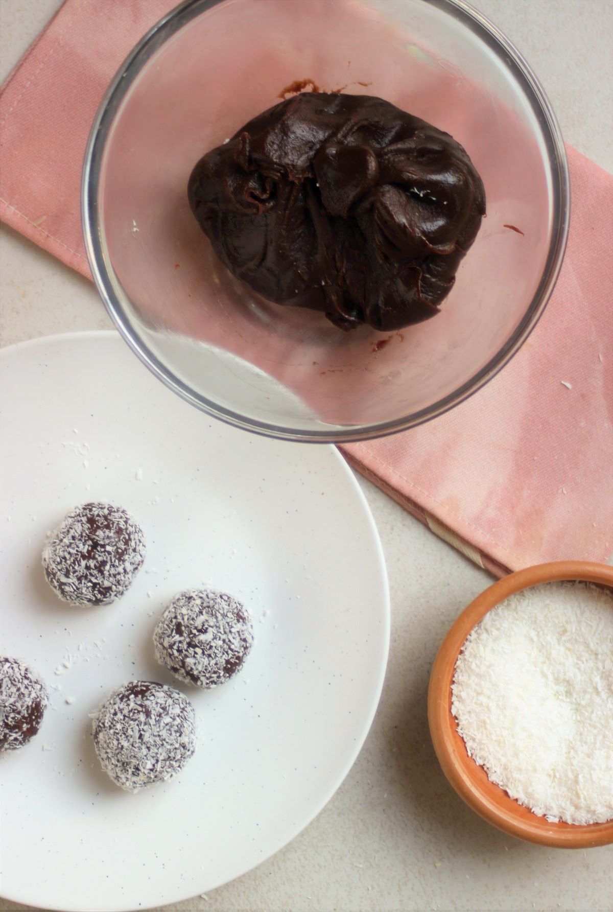 Glass bowl with chocolate batter, a white plate with brigadeiros rolled in coconut, and a bowl with shredded coconut.
