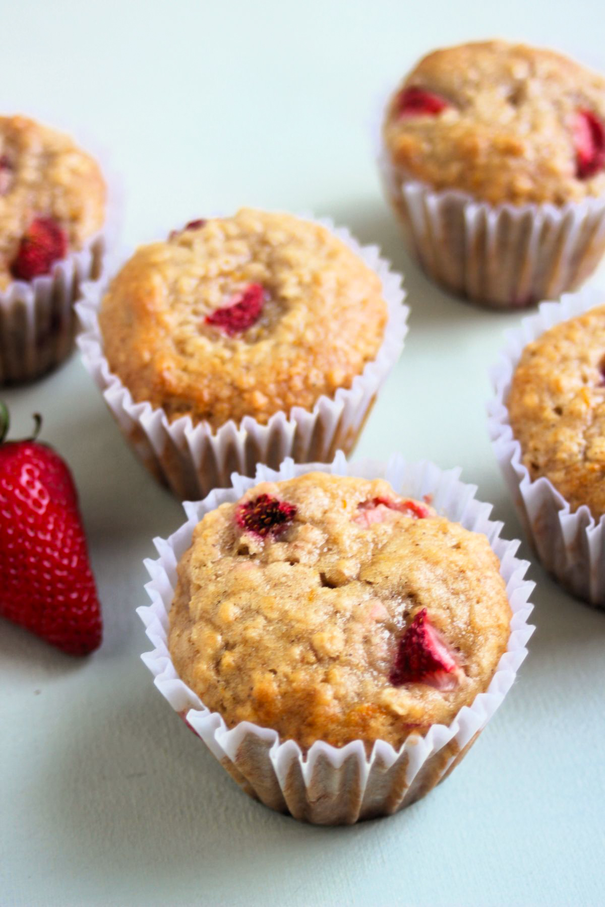 Strawberry oatmeal muffins with liners and a fresh strawberry on a sky light surface.