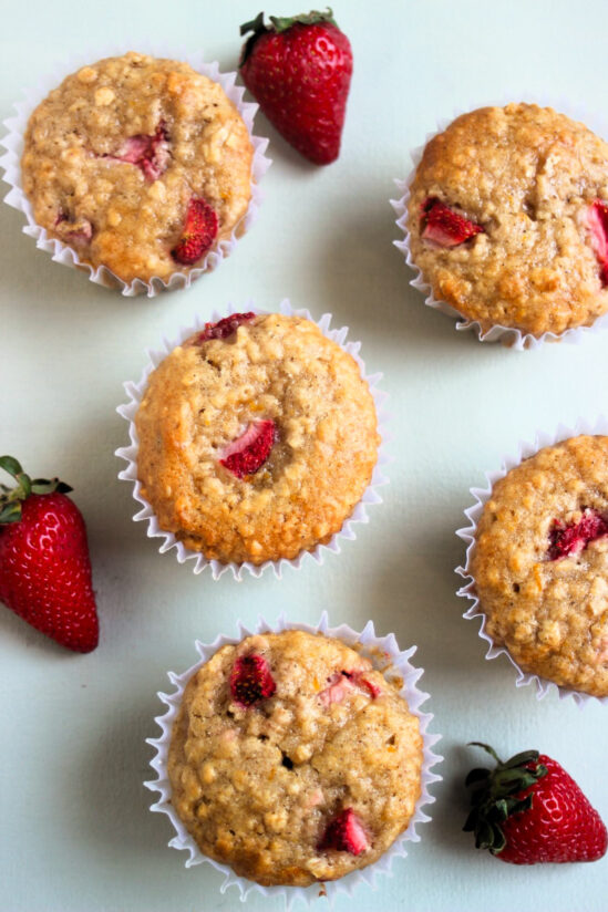 Strawberry oatmeal muffins with liners and fresh strawberries on a light blue surface seen from above.