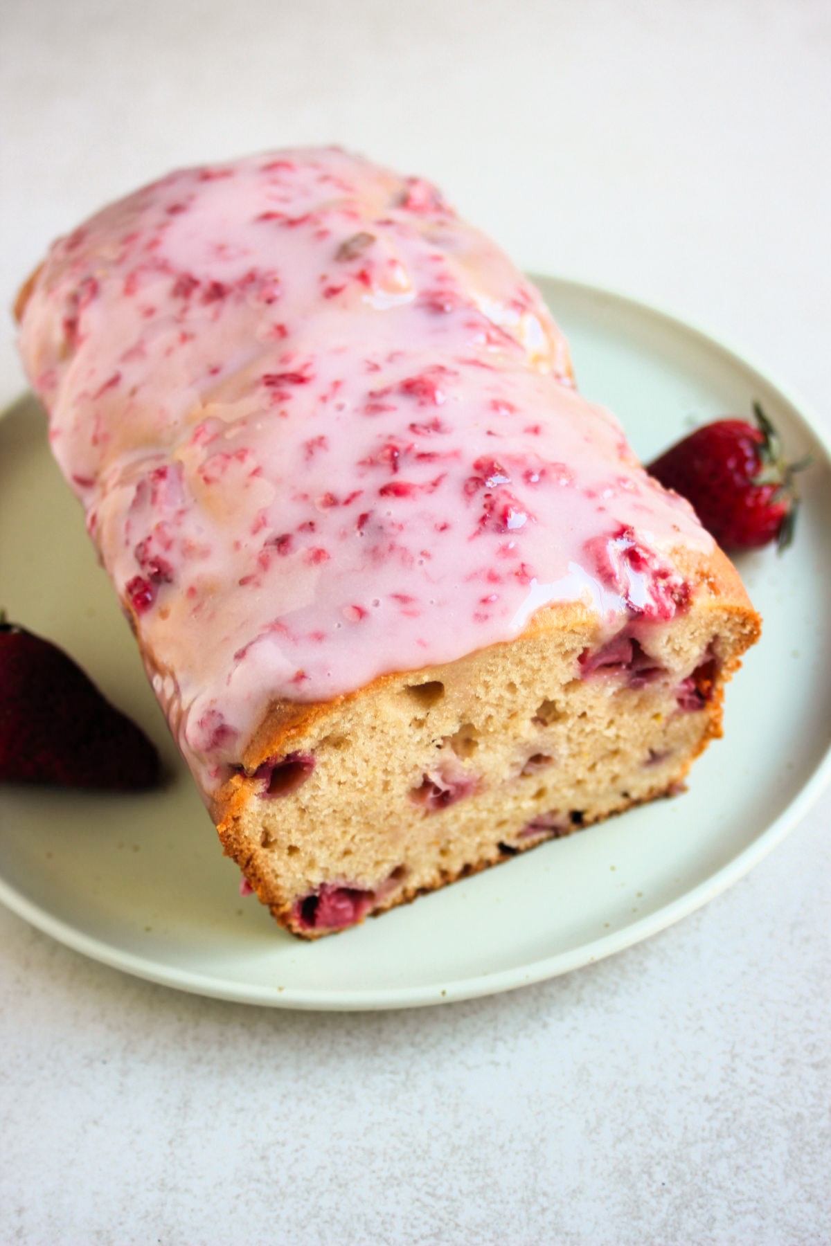 Strawberry bread with pink icing on an aqua-green plate. Fresh strawberries on the sides.