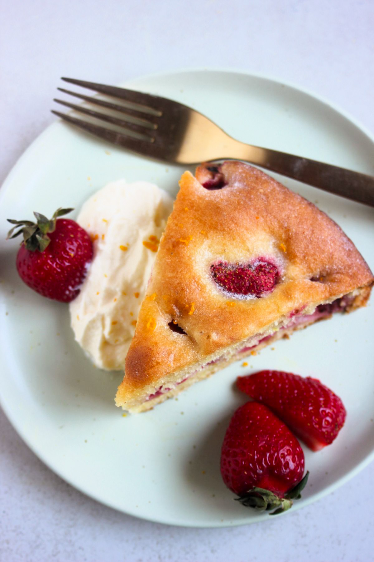 Top view of a portion of strawberry cake, a golden fork, whipped cream, and fresh strawberries on a pink plate.