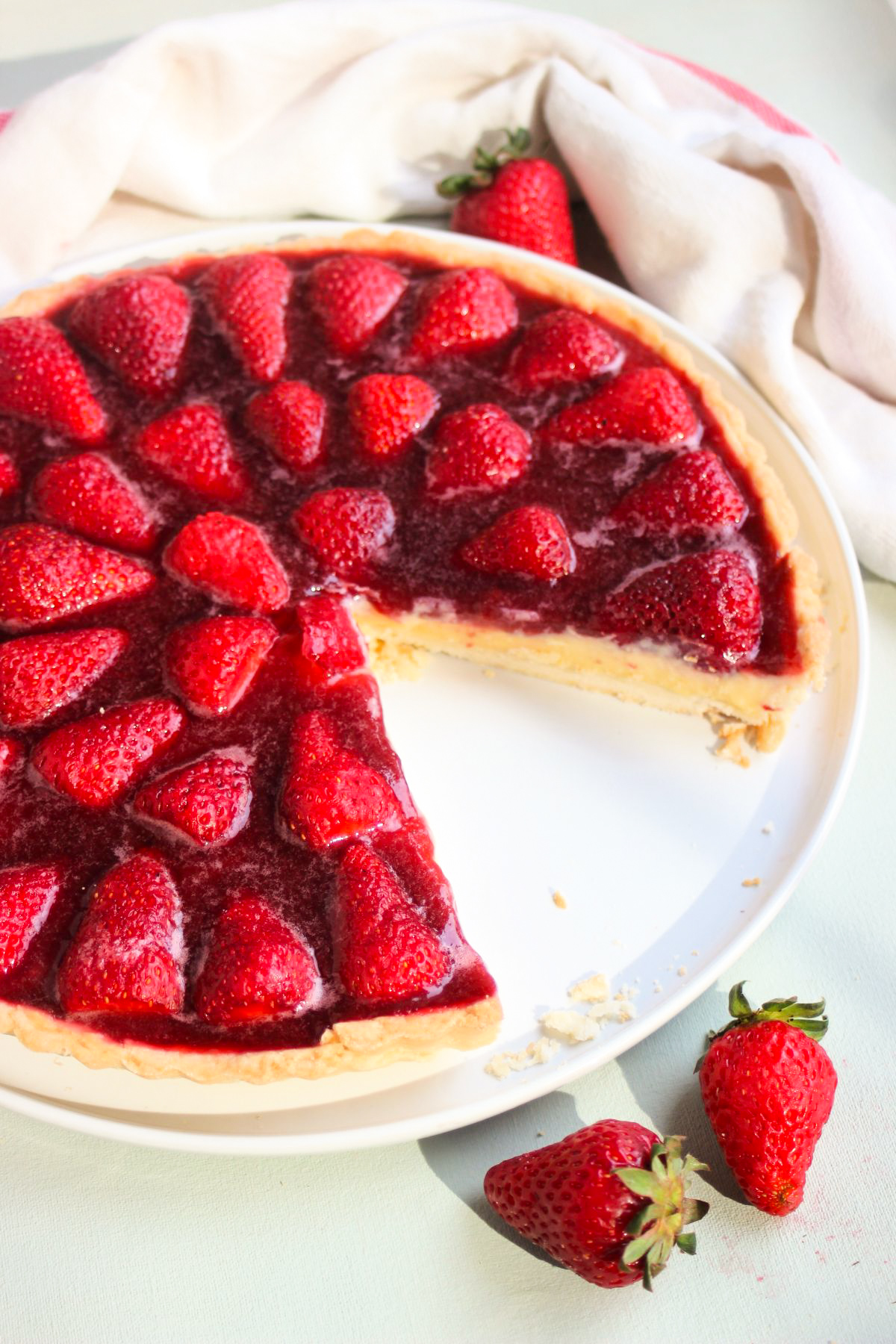 Partial strawberry tart on a white plate with a white kitchen towel and fresh strawberries. White surface.