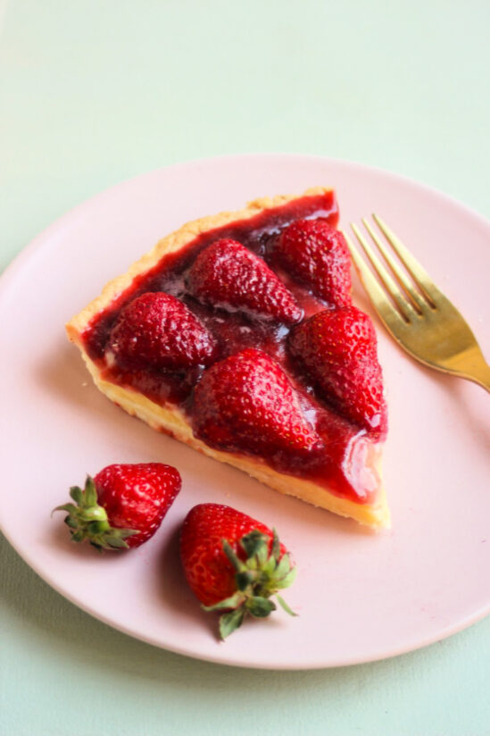 Slice of strawberry tart, two fresh strawberries, and a golden fork on a pink plate.