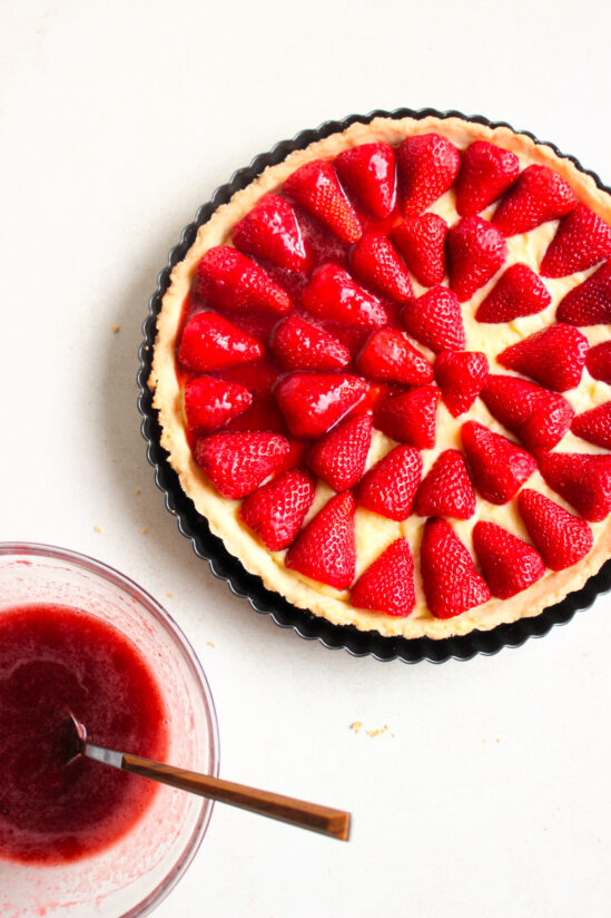 Top view of a strawberry tart in a round tart pan, before baking, and without the strawberry jelly sauce. A small bowl with the strawberry jelly sauce and a spoon.